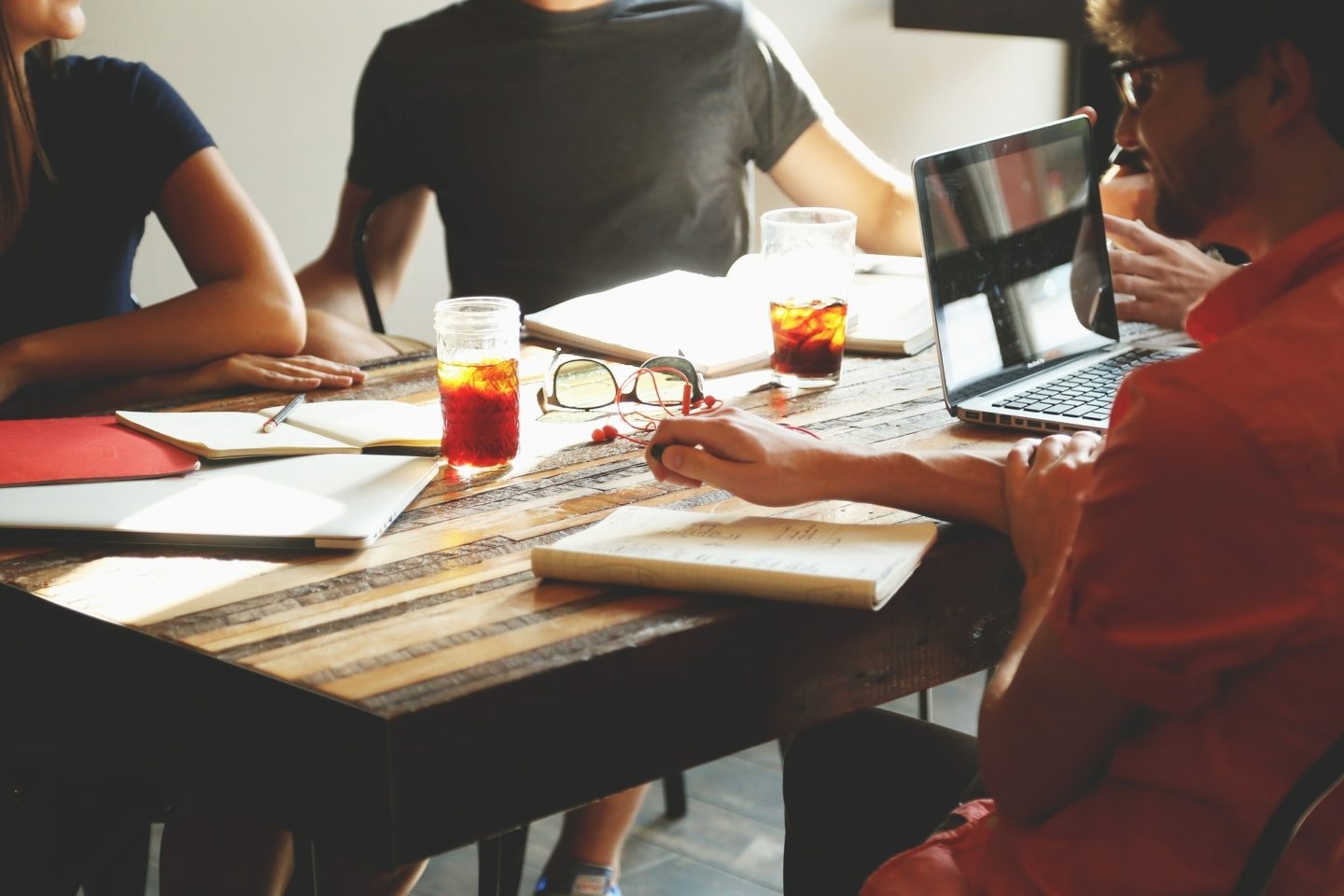 Four people sit at a wooden table. Each person has an open journal with notes. Two of the people have drinks in front of them, while one has an open laptop.