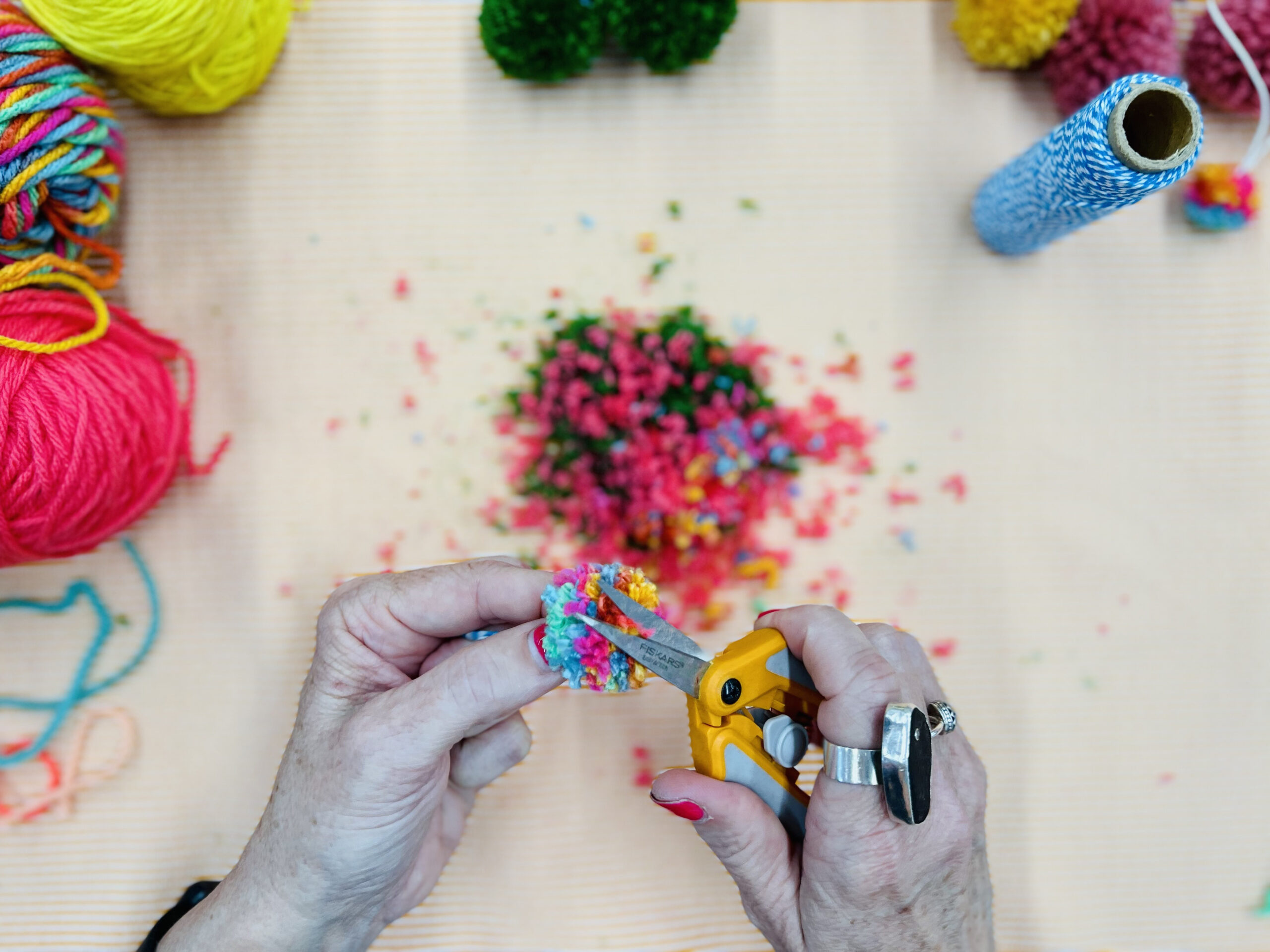 Adult Program image scaled An older woman's hands are holding a pair of scissors as she trims away a handmade yarn pom pom. Beneath her are the colorful bits of yarn trimmed away, and various balls of yarn used to make her craft.