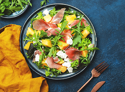 Delicious summer fresh salad with cantaloupe melon, prosciutto, soft cheese and arugula on blue table background, top view, copy space