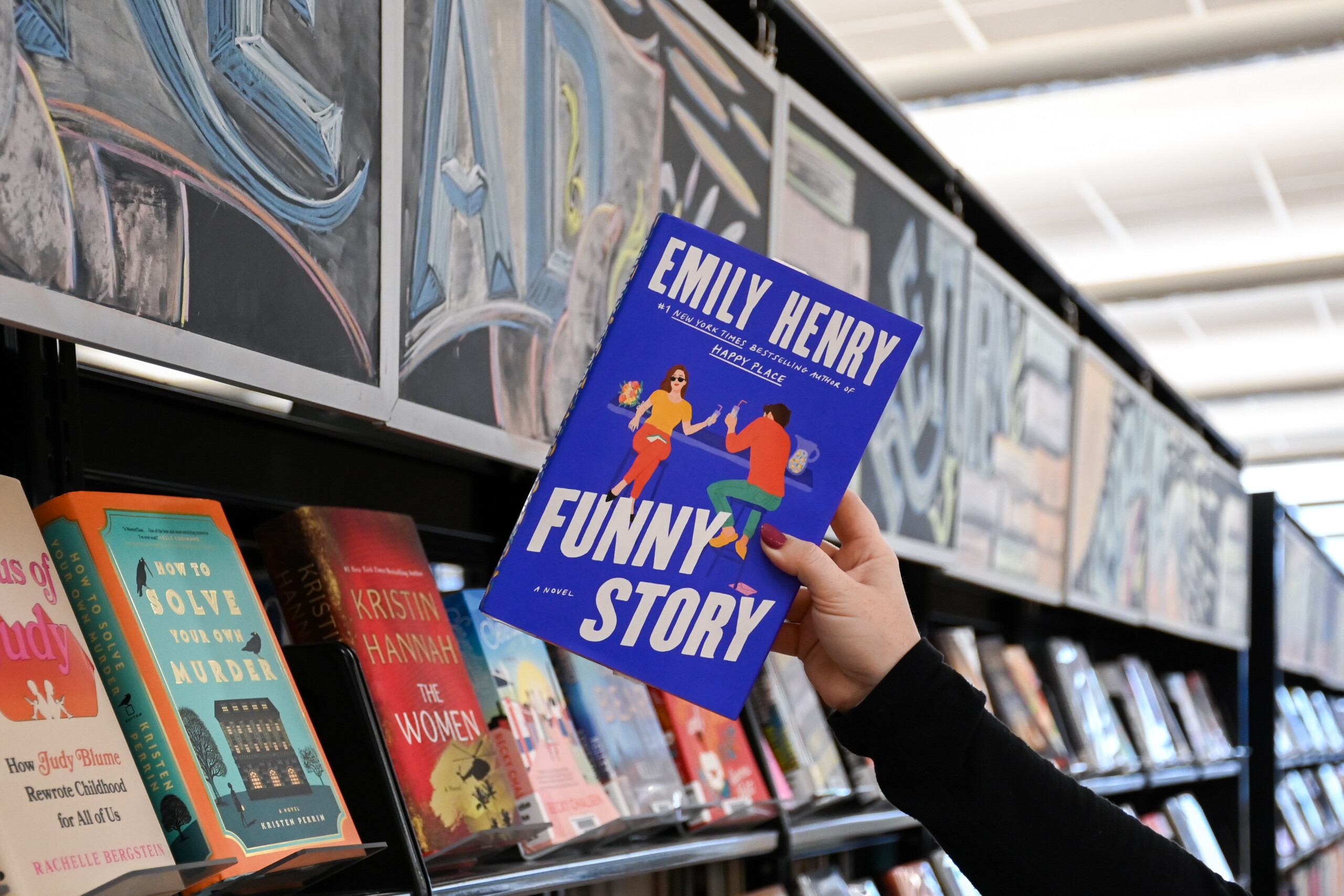 A person is holding the book "Funny Story" in front of bookshelf filled with books.