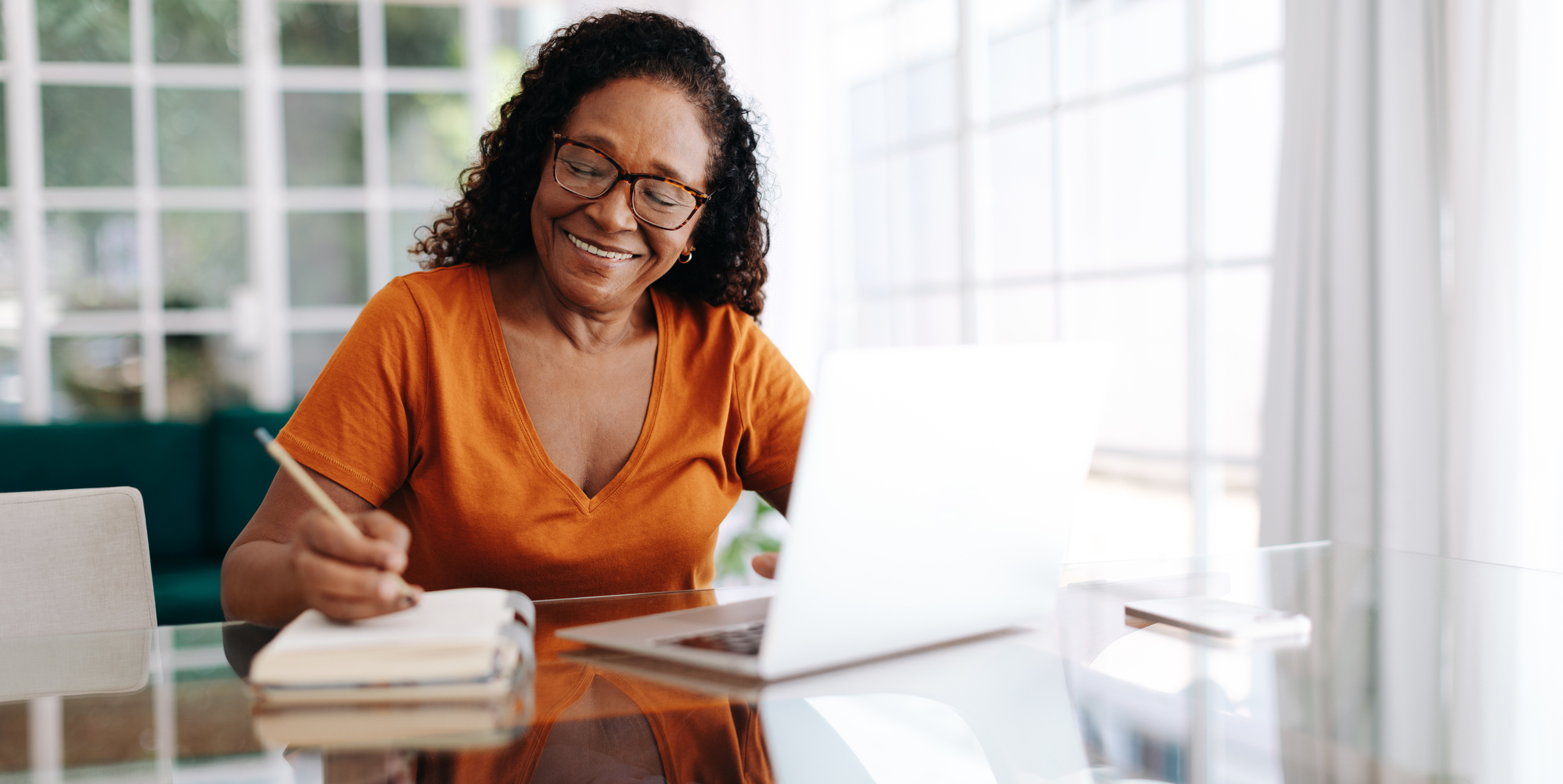 A dark-skinned woman with glasses is writing in a journal while working in front of her laptop.