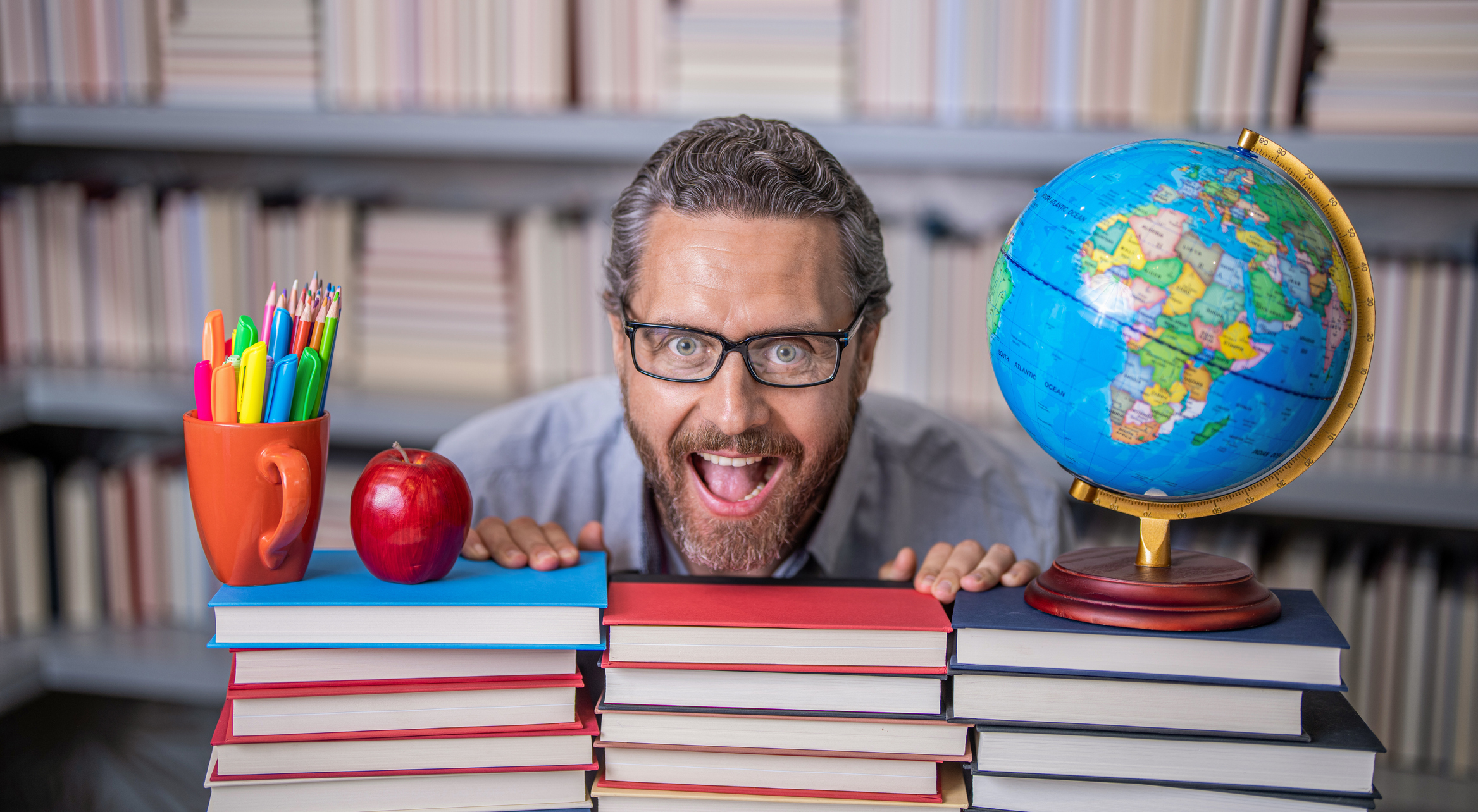 A teacher smiling by a stack of books and school supplies.