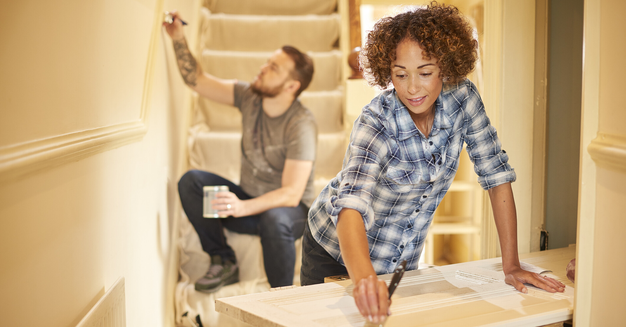 A young couple doing up their hallway . The man is painting the demo rail on the stairs covered in a dust sheet whilst his wife is painting a period door on a workbench .