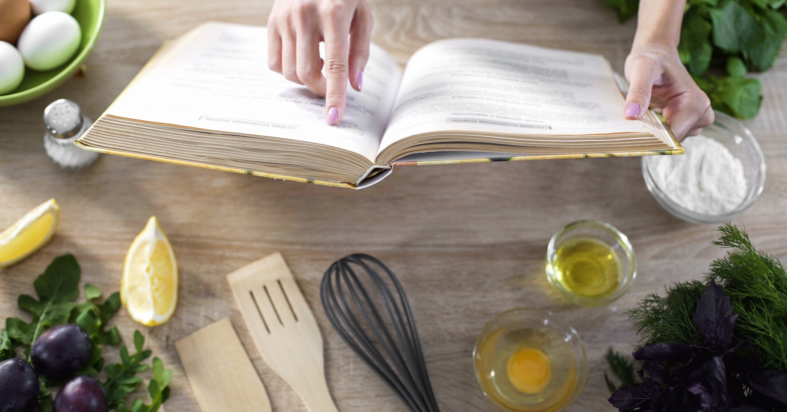 Person reads cookbook on table with kitchenware and ingredients.